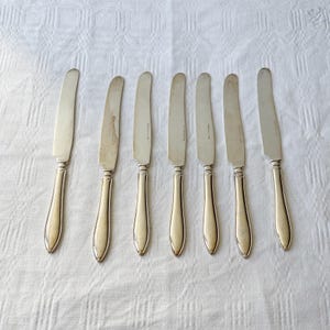 Może przedstawiać: Siedem srebrnych noży do masła ułożonych na białym obrusie. Noże mają zakrzywione ostrze i zaokrąglony uchwyt. Niektóre wykazują ślady zużycia. Noże są prawdopodobnie zabytkowe lub antyczne.