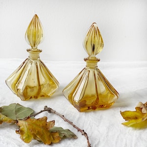 May include: Two amber glass perfume bottles with a fluted design and a pointed top. The bottles are sitting on a white surface with fall leaves.