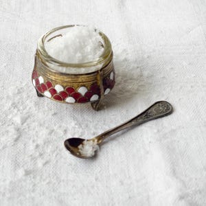May include: A small, ornate salt cellar filled with coarse salt, next to a small spoon. The salt cellar has a glass bowl and a decorative base with red, white, and gold accents. The spoon is silver-colored.
