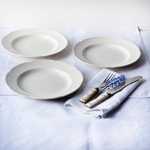 Puede incluir: Tres platos de cena de cerámica blanca con bordes festoneados están dispuestos sobre un mantel de lino blanco. Una servilleta de lino doblada con una flor azul, un tenedor y un cuchillo están sobre la mesa.