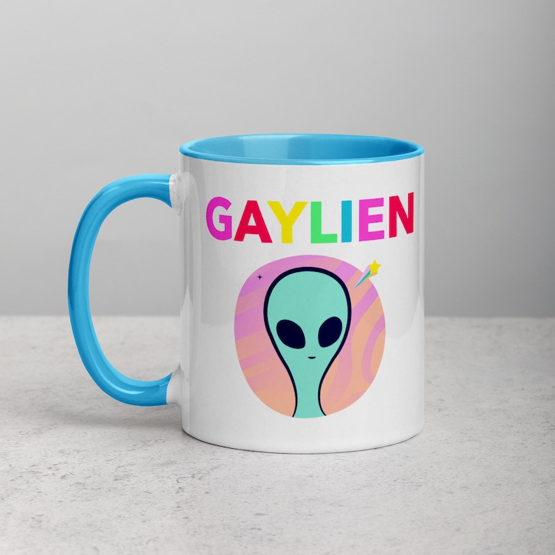 May include: A white ceramic mug with a light blue rim and handle. The mug features a colourful graphic of an alien with the text "GAYLIEN" in rainbow colours.