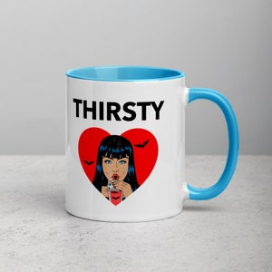 May include: White ceramic mug with a blue rim and handle. The mug has a black graphic of a woman with black hair drinking from a glass with a red heart and bats around it. The text "THIRSTY" is printed in black above the graphic.