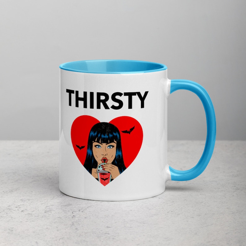 May include: White ceramic mug with a blue rim and handle. The mug has a black graphic of a woman with black hair drinking from a glass with a red heart and bats around it. The text "THIRSTY" is printed in black above the graphic.