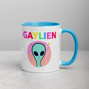 May include: White ceramic mug with a light blue rim and handle. The mug features a colourful graphic of an alien with the text "GAYLIEN" in rainbow colours.