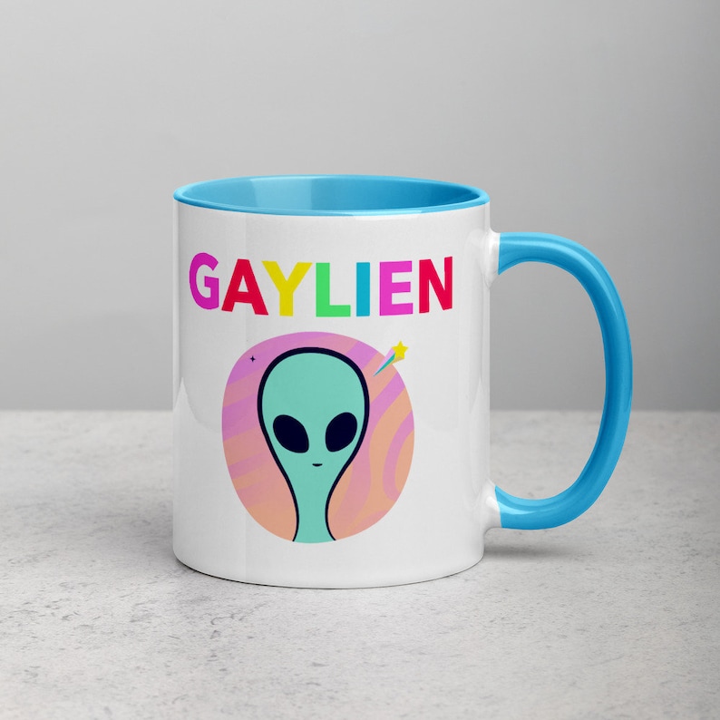 May include: White ceramic mug with a light blue rim and handle. The mug features a colourful graphic of an alien with the text "GAYLIEN" in rainbow colours.