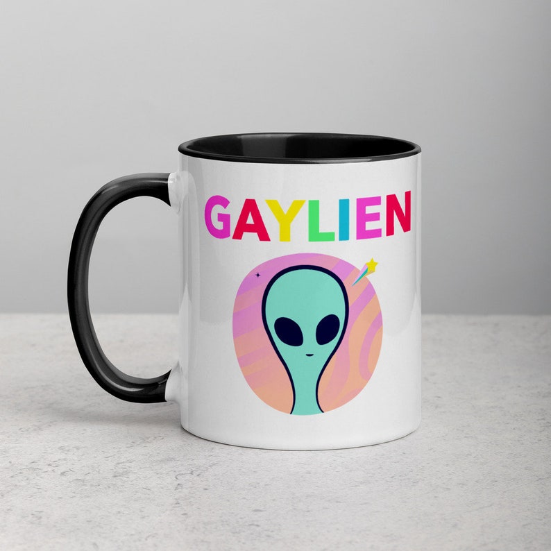 May include: White ceramic coffee mug with a black rim and handle. The mug features a colourful graphic of an alien with the text "GAYLIEN" in rainbow colours.