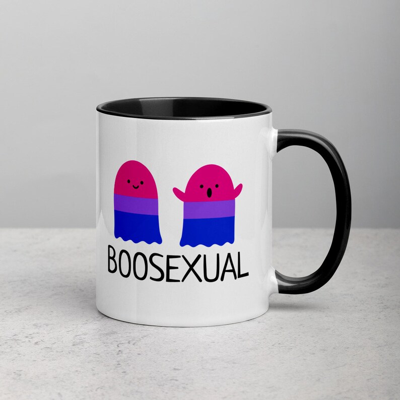 May include: White ceramic mug with a black rim and handle. The mug features two cartoon ghosts with the bisexual pride flag colors. The text "BOOSEXUAL" is printed below the ghosts.