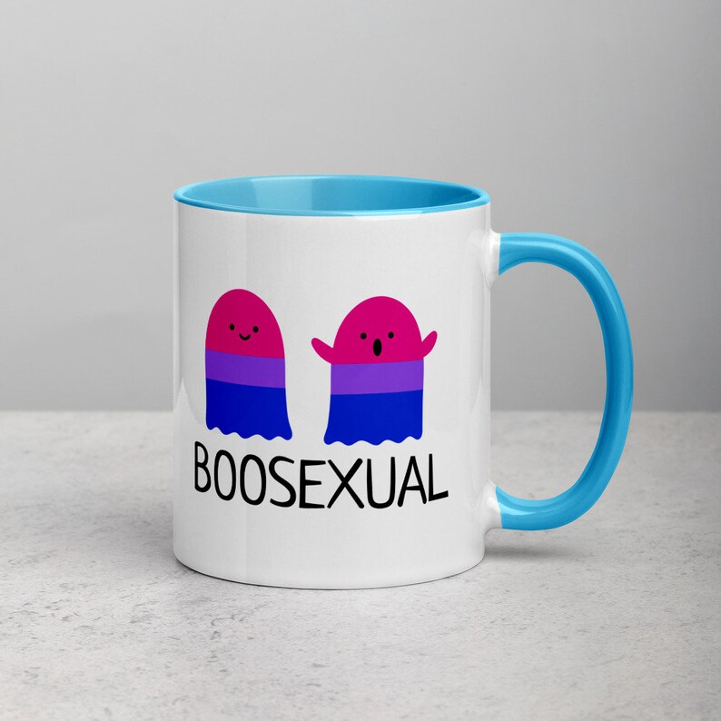 May include: A white ceramic mug with a light blue rim and handle. The mug features two cartoon ghosts with the bisexual pride flag colors. The text "BOOSEXUAL" is printed below the ghosts.