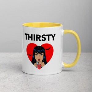 May include: White ceramic mug with a yellow rim and handle. The mug features a black and red graphic of a woman with black hair drinking from a glass with a straw. The text "THIRSTY" is printed in black above the graphic.