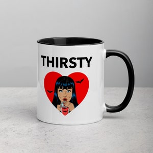 May include: White ceramic mug with a black rim and handle. The mug features a red heart with a black outline and a cartoon image of a woman with black hair drinking from a glass. The text "THIRSTY" is printed in black above the heart.