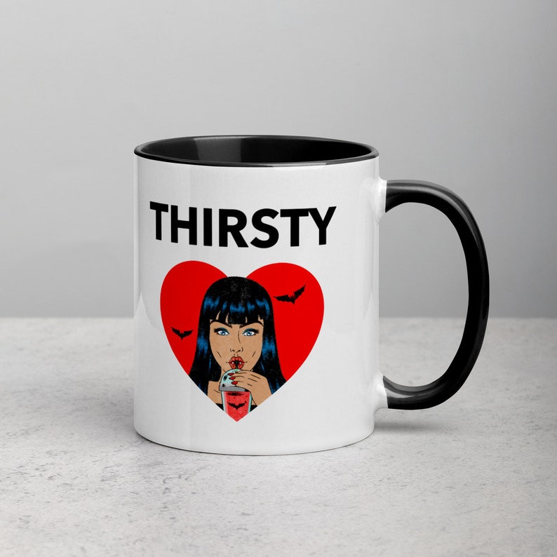 May include: White ceramic mug with a black rim and handle. The mug features a red heart with a black outline and a cartoon image of a woman with black hair drinking from a glass. The text "THIRSTY" is printed in black above the heart.