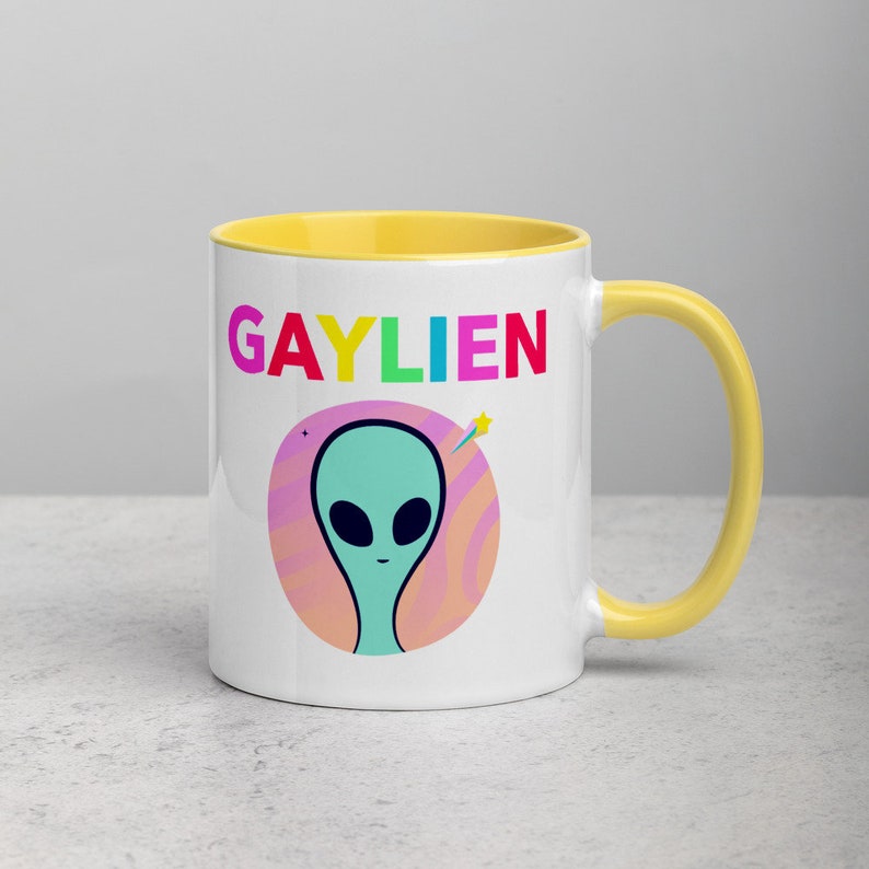 May include: White ceramic mug with a yellow rim and handle. The mug features a rainbow coloured word "GAYLIEN" and a cartoon alien with a pink and orange background.