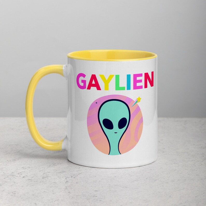 May include: White ceramic mug with a yellow rim. The mug features a colourful graphic of an alien with the text "GAYLIEN" in rainbow colours.