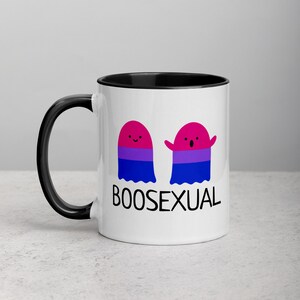 May include: White ceramic mug with a black rim and handle. The mug features two cartoon ghosts with the bisexual pride flag colors. The text "BOOSEXUAL" is printed below the ghosts.
