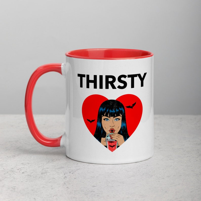 May include: White ceramic mug with a red rim and handle. The mug features a black and red graphic of a woman with black hair drinking from a glass with a red liquid. The text "THIRSTY" is printed in black above the graphic.
