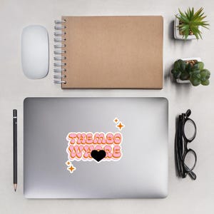 Pode incluir: Um autocolante com o texto "THEMBO WHERE" em letras de bolhas laranja e rosa, com um coração preto e detalhes de estrelas laranja. O autocolante está num portátil, com uma caneta, um caderno, um rato e pequenas plantas por perto.