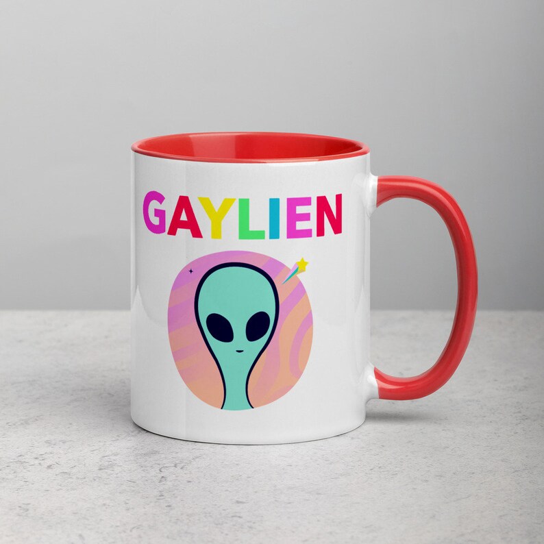 May include: White ceramic mug with a red rim and handle. The mug features a graphic of a green alien with a rainbow coloured word "GAYLIEN" above it.
