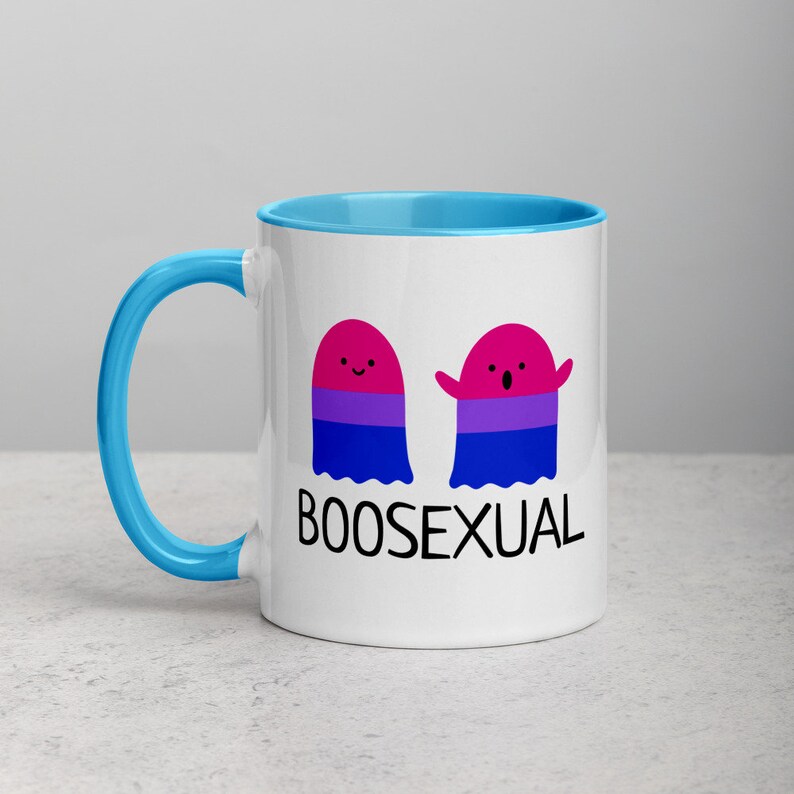 May include: White ceramic mug with a blue rim and handle. The mug features two cartoon ghosts with the bisexual pride flag colors. The text "BOOSEXUAL" is printed below the ghosts.