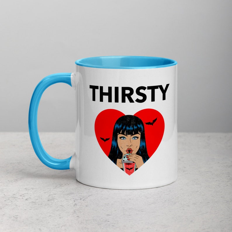 May include: White ceramic mug with a blue rim. The mug features a black and red graphic of a woman with black hair drinking from a glass with a straw. The text "THIRSTY" is printed in black above the graphic.