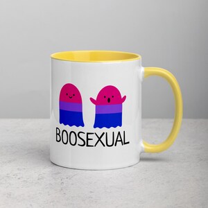 May include: White ceramic mug with a yellow rim and handle. The mug features two cartoon ghosts with a bisexual pride flag design and the text "BOOSEXUAL".