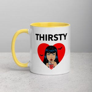 May include: White ceramic mug with a yellow rim. The mug features a black and red graphic of a woman with black hair drinking from a red cup. The text "THIRSTY" is printed in black above the graphic.