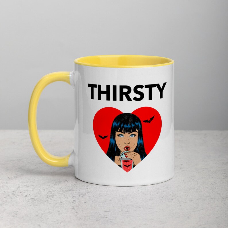 May include: White ceramic mug with a yellow rim. The mug features a black and red graphic of a woman with black hair drinking from a red cup. The text "THIRSTY" is printed in black above the graphic.