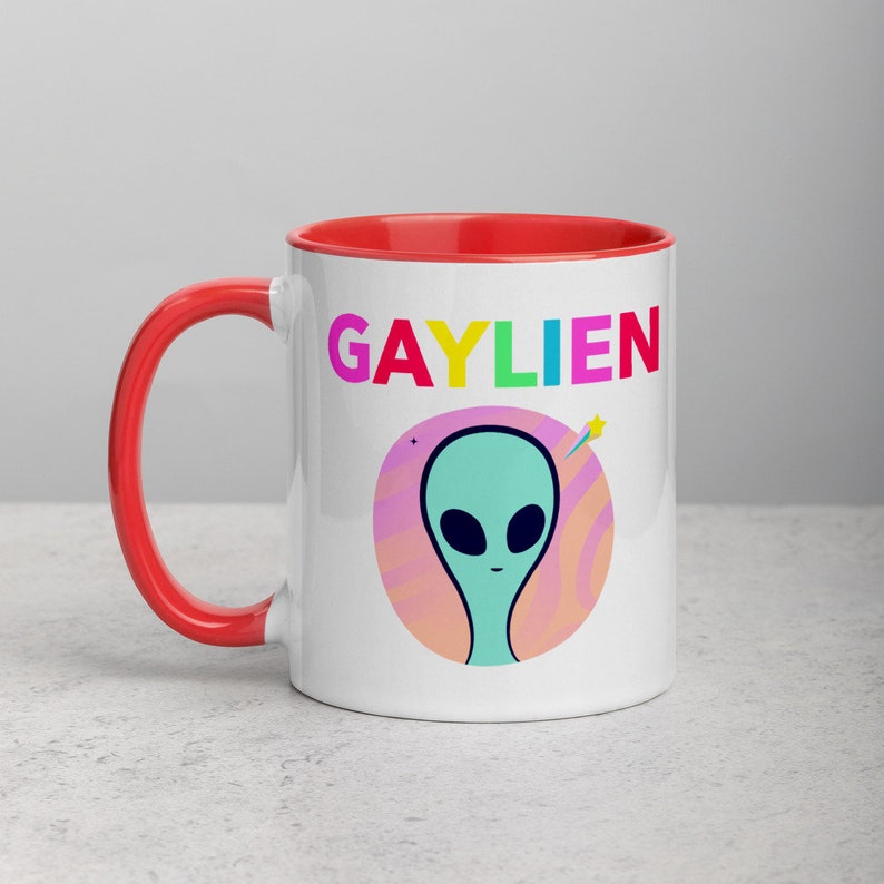 May include: White ceramic mug with a red rim. The mug features a graphic of a green alien with a rainbow coloured word "GAYLIEN" above it.