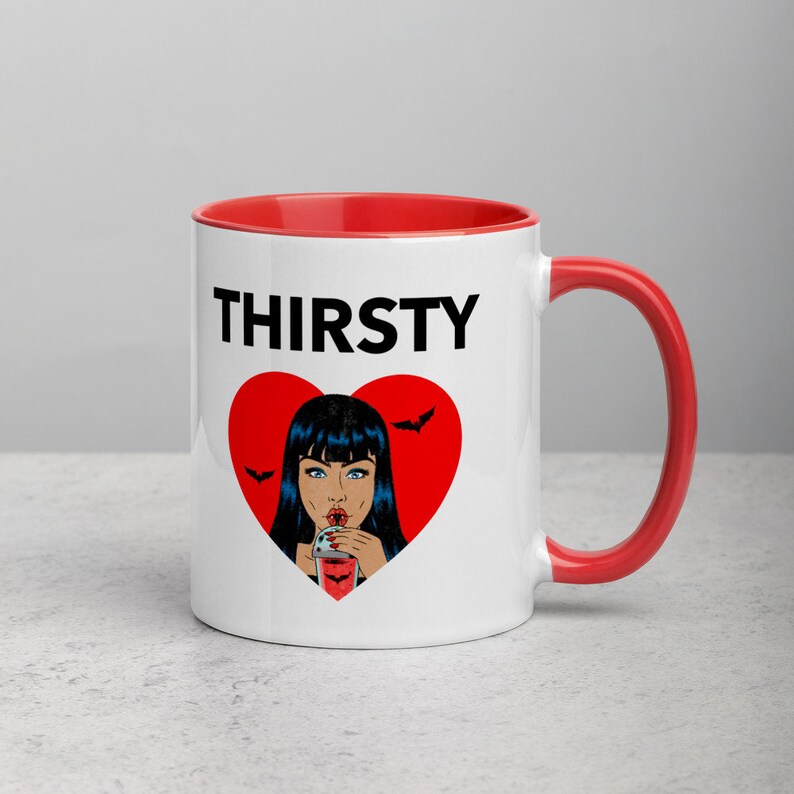 May include: White ceramic mug with a red rim and handle. The mug has a black graphic of a woman with black hair drinking from a glass with a red heart and bats around it. The text "THIRSTY" is printed in black above the graphic.
