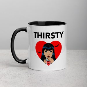 May include: White ceramic mug with a black rim and handle. The mug features a red heart with a black outline and a cartoon image of a woman with black hair drinking from a red cup. The text "THIRSTY" is printed in black above the heart.