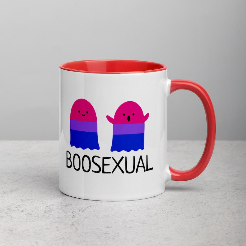 May include: White ceramic mug with a red rim and handle. The mug features two cartoon ghosts with the bisexual pride flag colors. The text "BOOSEXUAL" is printed below the ghosts.