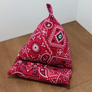 May include: A red bandana-patterned phone stand. The stand is triangular, with a folded base and a raised, peaked top. The fabric features a classic paisley design in white against a red background. The stand is designed to prop up a phone or tablet.