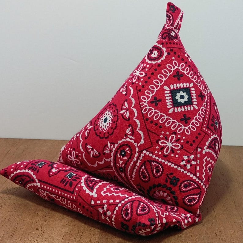 May include: A red bandana-patterned book stand and a matching bolster pillow. The fabric features a white paisley and floral design. The book stand is triangular, and the bolster is cylindrical.
