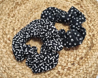 May include: Two black and white scrunchies. One features a bone pattern, the other a paw print design. The scrunchies are made of fabric and are designed to hold hair in place. They are placed on a woven, tan surface.