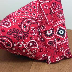 May include: A red bandana-patterned fabric item, featuring a classic paisley design with white and black accents. The fabric is folded and sewn, showcasing the intricate pattern and texture. The item is likely a small bag or accessory.