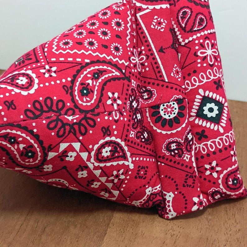 May include: A red bandana-patterned fabric item, featuring a classic paisley design with white and black accents. The fabric is folded and sewn, showcasing the intricate pattern and texture. The item is likely a small bag or accessory.