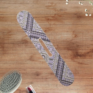 May include: A long, narrow, flat hair accessory with a rounded end and a central elongated cutout. The surface features a repeating geometric pattern in shades of purple, black, and white, consisting of intricate lines, squares, and zigzags.