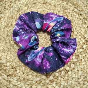 May include: A scrunchie with a vibrant galaxy print featuring swirling purples, blues, and pinks. The fabric design includes planets and stars, creating a cosmic theme. The scrunchie is made of a soft, gathered material.