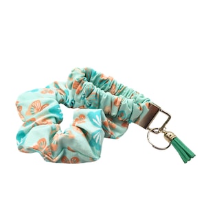 May include: A light teal scrunchie and wristlet set with a floral pattern in orange and teal. The wristlet has a silver-tone metal clasp, a small gold-tone charm, and a green tassel.