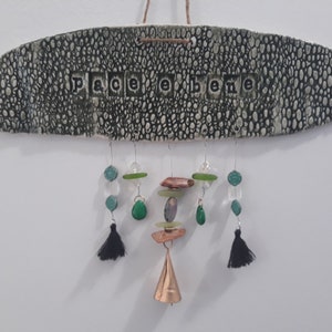 May include: A green ceramic oval sign with the words "Peace & Belle" in black lettering. The sign has a hanging mobile with green, brown, and clear glass beads, a copper bell, and black tassels.