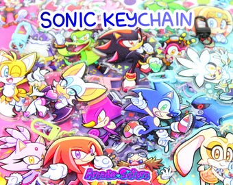 Może przedstawiać: Kolekcja kolorowych breloków z postaciami z Sonic the Hedgehog. Breloki przedstawiają różne postacie, takie jak Sonic, Tails, Knuckles i Shadow. Tekst "SONIC KEYCHAIN" jest wyświetlany u góry. Logo Arcade Sisters znajduje się na dole.
