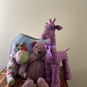 Op de afbeelding: Drie handgemaakte knuffels: een nijlpaard, een teddybeer en een giraf. Het nijlpaard en de giraf zijn bedekt met een bloemenstof met rode, roze en blauwe bloemen. De teddybeer is bruin met verticale strepen. De giraf heeft een paars lint.