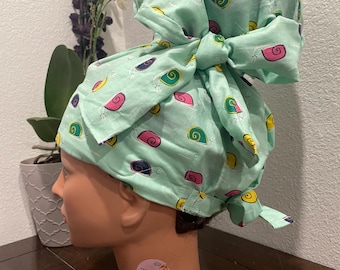 May include: A light green surgical scrub cap with a bow, patterned with colorful snails. The cap is tied in a bow on top of the head. The snails are in shades of pink, yellow, blue, and green.