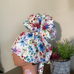 May include: A white surgical scrub hat with a floral print featuring pink, blue, green, and red flowers. The hat is tied in a bun on top of the head. The hat has a long tie that is tied in a bow at the back of the neck. The hat is on a mannequin head. The hat has a small pink sticker with the text "Thank you" on it.