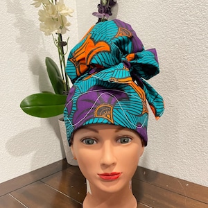 May include: A patterned scrub cap in teal, orange, and purple, with a bow on top. The cap features a repeating floral design and is displayed on a mannequin head. The background includes a white wall and a wooden surface.