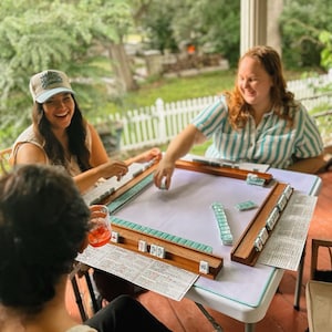 May include: Three people are gathered around a white table playing Mahjong. The game set includes wooden racks, tiles, and score sheets. The players are smiling and laughing, enjoying the outdoor setting.