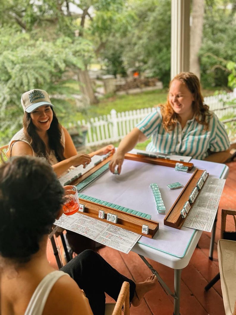 May include: Three people are gathered around a white table playing Mahjong. The game set includes wooden racks, tiles, and score sheets. The players are smiling and laughing, enjoying the outdoor setting.