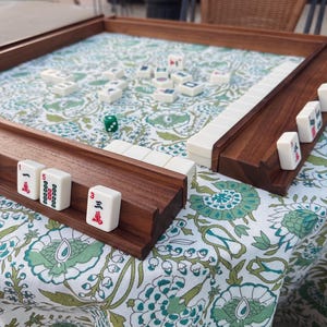 May include: A wooden mahjong game set featuring a square board with a raised frame and two wooden tile racks. The board surface is covered in a white fabric with a green and teal floral pattern. White mahjong tiles with red and black markings are arranged on the racks and scattered across the board, accompanied by a single green die.