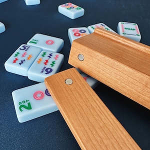 May include: A light blue Mahjong tile set with wooden tile holders. The tiles have colorful symbols and numbers. The wooden holders are rectangular and have magnets. The tiles are scattered on a dark blue surface.