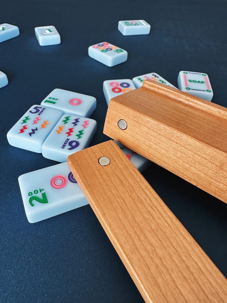 May include: A light blue Mahjong tile set with wooden tile holders. The tiles have colorful symbols and numbers. The wooden holders are rectangular and have magnets. The tiles are scattered on a dark blue surface.