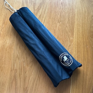 May include: A navy blue, cylindrical item, possibly a rolled-up umbrella or similar product, is shown. The item is secured with a white drawstring. The Backyard Yacht Club logo is visible on the side.